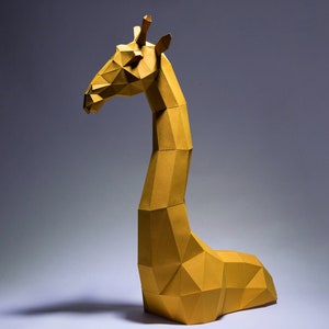 Op de afbeelding: Een gele origami-giraffenbeeld met een geometrisch ontwerp. De giraffe staat op een wit oppervlak met een grijze achtergrond.