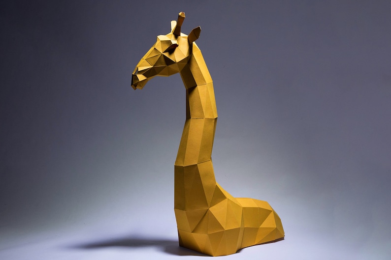 Op de afbeelding: Een gele origami-giraffenbeeld met een geometrisch ontwerp. De giraffe staat op een wit oppervlak met een grijze achtergrond.