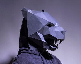 Pode incluir: Uma máscara de papel maché cinza em forma de urso com uma expressão feroz. A máscara é feita de papel dobrado e tem um design geométrico.