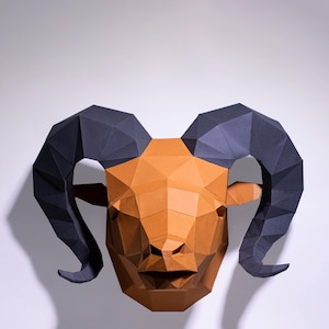 Op de afbeelding: Een papieren sculptuur van een rammenkop met bruin en zwart papier. De rammenhoorns zijn gekruld en wijzen omhoog. Het gezicht van de ram is gedetailleerd met een neus, mond en ogen.