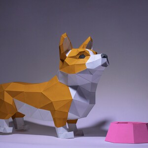 K&ouml;nnte beinhalten: Ein Papiermodell eines Corgi-Hundes, das neben einer rosa Hundenapf steht. Der Hund ist aus wei&szlig;em und orangefarbenem Papier gefertigt und hat ein realistisches Aussehen.