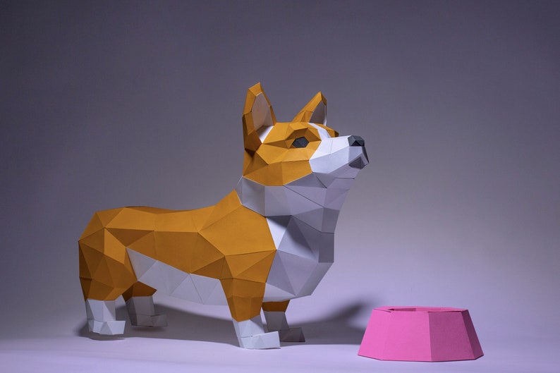 K&ouml;nnte beinhalten: Ein Papiermodell eines Corgi-Hundes, das neben einer rosa Hundenapf steht. Der Hund ist aus wei&szlig;em und orangefarbenem Papier gefertigt und hat ein realistisches Aussehen.