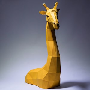 Op de afbeelding: Een gele papieren giraffesculptuur met een geometrisch ontwerp. De giraffe zit op een wit oppervlak met een grijze achtergrond.