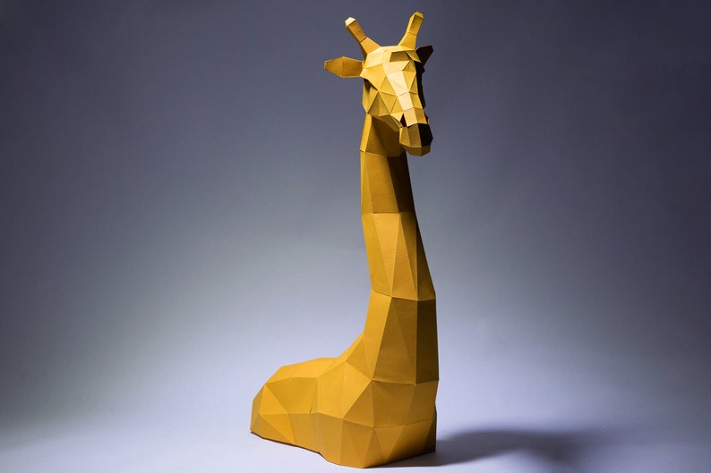 Op de afbeelding: Een gele papieren giraffesculptuur met een geometrisch ontwerp. De giraffe zit op een wit oppervlak met een grijze achtergrond.