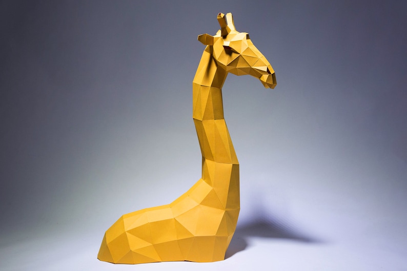 Op de afbeelding: Een gele origami-giraf-sculptuur met een geometrisch ontwerp. De giraf zit op een wit oppervlak.