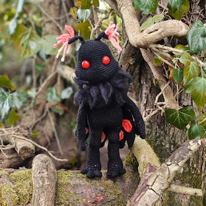Puede incluir: Una muñeca de ganchillo negra hecha a mano con ojos rojos y detalles de alas rojas y negras. La polilla tiene antenas rosas y un cuello negro esponjoso. La muñeca está de pie en una rama de árbol.