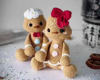 May include: Two crocheted gingerbread people, one with a red bow and the other with a white and blue bow, sitting on a white surface with sprinkles.