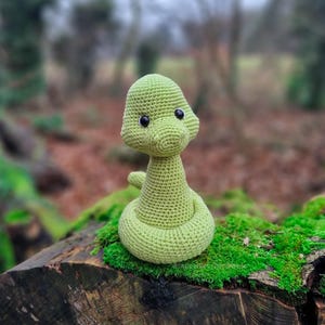 May include: A light green crocheted snake toy with a coiled body and a rounded head. The snake features two small, round, black plastic eyes. The texture of the yarn is visible throughout the entire surface of the stuffed animal.