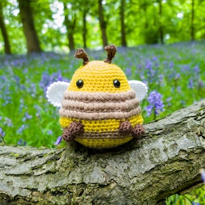 May include: A crocheted bee toy with yellow and brown stripes, white wings, and black eyes. The bee is sitting on a tree branch in a woodland setting.