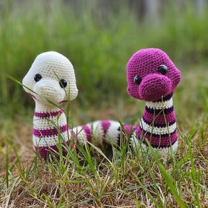May include: Two handmade crochet snake toys with large black eyes rest in the grass. One is white with purple stripes, and the other is purple with white and purple stripes. The toys have a whimsical, cartoonish appearance.