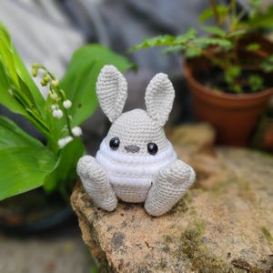 May include: A handmade crochet bunny in shades of gray and white. The bunny has large, upright ears, black eyes, and a small gray nose. It is sitting on a rock, with a blurred background of greenery and a potted plant.