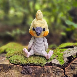 Puede incluir: Un juguete de pájaro cacatúa tejido a crochet con hilo amarillo, naranja y gris. El pájaro está sentado en un tronco cubierto de musgo.