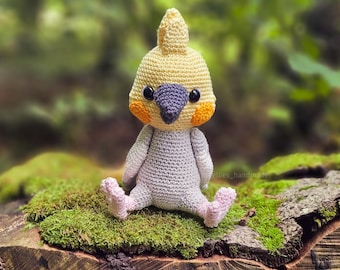 May include: A crocheted cockatiel bird toy with yellow, orange, and gray yarn. The bird is sitting on a mossy log.