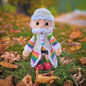 May include: A handmade crochet doll of a bearded figure wearing a cream-colored coat with rainbow trim and sleeves. The doll has a matching hat and is standing on green grass with fallen leaves. The doll's shoes are red.