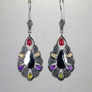 May include: A pair of silver dangle earrings with a teardrop-shaped design. The earrings feature a variety of colorful gemstones, including red, white, yellow, green, and purple.