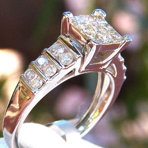 May include: A silver ring with a large square-shaped diamond in the center and smaller diamonds set in a band around the center stone.