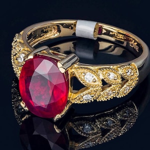May include: A gold ring with a large, oval-cut ruby gemstone as the centerpiece. The ring features intricate gold detailing with small, sparkling diamonds and a reflective surface.