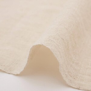 Puede incluir: Primer plano de un trozo de tela de lino de color natural doblado. La tela tiene un tejido ligeramente texturizado y un tono suave y neutro, con sutiles variaciones de color. Los bordes de la tela son visibles, mostrando el grosor y la textura del material.