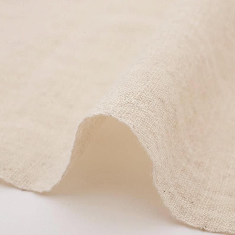 Puede incluir: Primer plano de un trozo de tela de lino de color natural doblado. La tela tiene un tejido ligeramente texturizado y un tono suave y neutro, con sutiles variaciones de color. Los bordes de la tela son visibles, mostrando el grosor y la textura del material.
