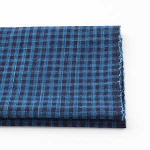 May include: Folded blue and navy gingham fabric. The fabric features a classic checkered pattern, with a frayed edge visible on the right side. The fabric is folded over itself, showing multiple layers.