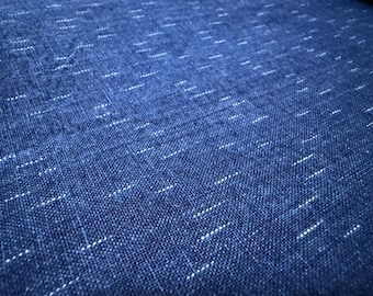 May include: Close-up of a textured, indigo blue fabric with a subtle pattern of white dashes. The weave is visible, creating a tactile appearance. The fabric has a slightly faded look, suggesting a vintage or well-worn aesthetic.