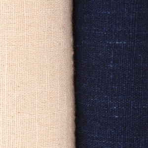 Puede incluir: Primer plano de dos telas enrolladas. Una es de color blanco roto natural, y la otra es azul marino oscuro. Ambas telas tienen una apariencia texturizada y tejida, lo que sugiere lino o un material similar. La imagen resalta el contraste de color y textura.