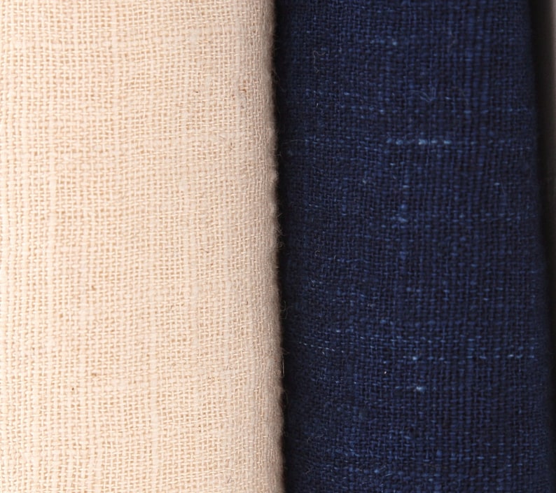 Puede incluir: Primer plano de dos telas enrolladas. Una es de color blanco roto natural, y la otra es azul marino oscuro. Ambas telas tienen una apariencia texturizada y tejida, lo que sugiere lino o un material similar. La imagen resalta el contraste de color y textura.