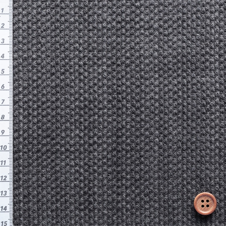 May include: Close-up of a dark grey fabric with a textured, woven pattern. A wooden button with four holes is in the lower right corner. A ruler with inch markings is on the left side of the fabric.