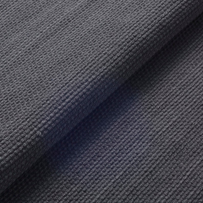 May include: Close-up of a folded section of dark grey fabric. The material has a textured, waffle-like weave, creating a grid pattern across the surface. The fabric is tightly woven, with a slightly uneven texture.