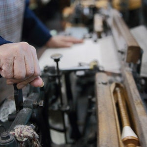 May include: A close-up of a person's hand operating a vintage loom. The loom is made of wood and metal and has a spool of thread on the right side. The person's hand is gripping a lever on the loom.