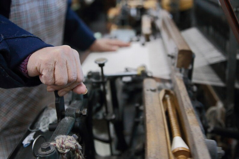 May include: A close-up of a person's hand operating a vintage loom. The loom is made of wood and metal and has a spool of thread on the right side. The person's hand is gripping a lever on the loom.
