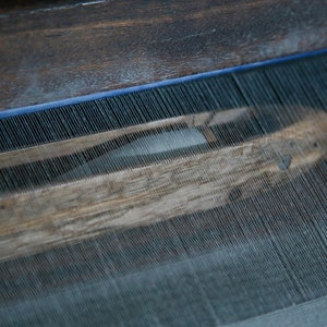 May include: Close-up of a wooden loom with threads woven through it. The threads are white and the loom is made of dark wood.
