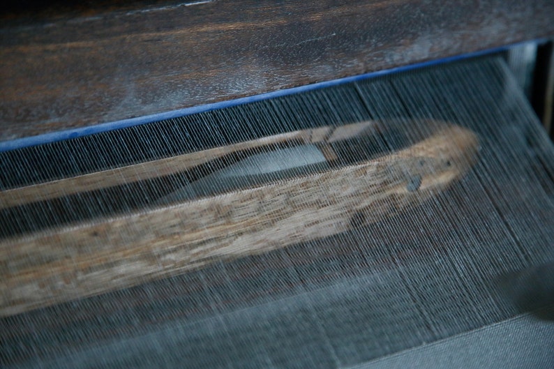 May include: Close-up of a wooden loom with threads woven through it. The threads are white and the loom is made of dark wood.