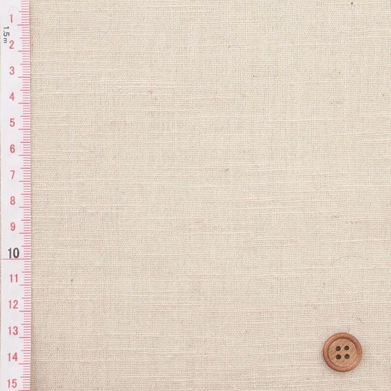 Puede incluir: Primer plano de tela de lino beige con un bot&oacute;n de madera. Una regla con medidas m&eacute;tricas est&aacute; a la izquierda. La tela tiene una textura sutil, ideal para costura.