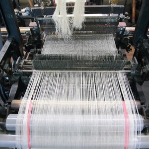 May include: A close-up of a vintage loom with white thread being woven. The loom is made of metal and has a wooden frame. The thread is being wound around a spool on the right side of the loom.