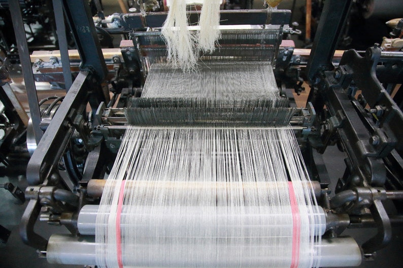 May include: A close-up of a vintage loom with white thread being woven. The loom is made of metal and has a wooden frame. The thread is being wound around a spool on the right side of the loom.
