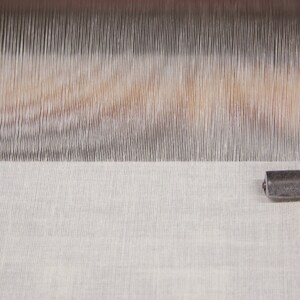 May include: Close-up of a white fabric being woven on a loom. The fabric is smooth and has a subtle texture. The loom is made of metal and wood.