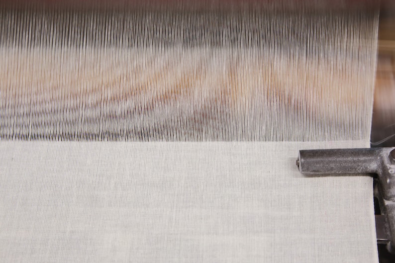 May include: Close-up of a white fabric being woven on a loom. The fabric is smooth and has a subtle texture. The loom is made of metal and wood.