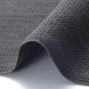 May include: A close-up shot of a folded, dark grey woven fabric. The textile has a textured, raised pattern, and the edges are neatly finished. The fabric appears soft and pliable, with a slight sheen.