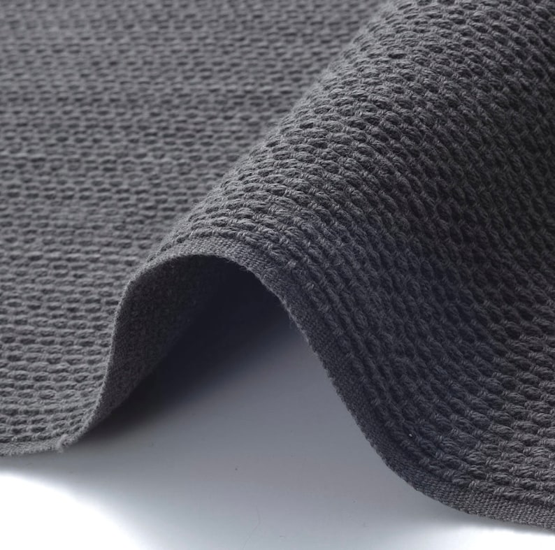 May include: A close-up shot of a folded, dark grey woven fabric. The textile has a textured, raised pattern, and the edges are neatly finished. The fabric appears soft and pliable, with a slight sheen.