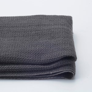 May include: A folded, dark grey woven textile with a textured, grid-like pattern. The fabric appears soft and pliable, with visible folds and a subtle sheen. The neutral colour and texture suggest a blanket or throw.