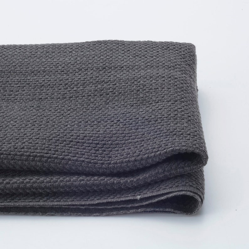 May include: A folded, dark grey woven textile with a textured, grid-like pattern. The fabric appears soft and pliable, with visible folds and a subtle sheen. The neutral colour and texture suggest a blanket or throw.