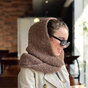 Könnte beinhalten: Ein brauner, strukturierter Kapuzenschal aus einem flauschigen, fleeceartigen Material, der sich um Kopf und Hals legt. Die Kapuze ist in das Design des Schals integriert und bietet eine vollständige Abdeckung für Kopf und Schultern.