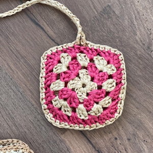 May include: A square, hand-crocheted coaster or small bag with a woven strap. The coaster features a floral pattern in shades of pink and beige, with a beige border. The background is a wooden surface.
