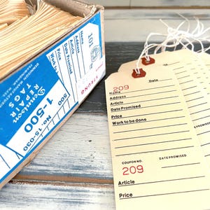 May include: A box of 500 blank yellow paper tags with string attached. The tags have lines for writing information, including a coupon number, date promised, article, and price. The box is blue and white with the text "1-500 Repair Tags" printed on it.