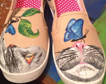 Könnte beinhalten: Ein Paar beigefarbene Slip-on-Schuhe aus Canvas mit handgemalten Designs. Ein Schuh zeigt einen Schmetterling, ein Blatt und ein Katzen Gesicht. Der andere Schuh zeigt einen Schmetterling und ein Katzen Gesicht. Beide Schuhe haben ein rosa-weißes Punktefutter.