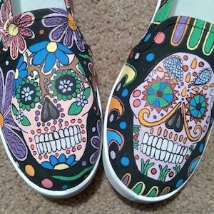 May include: A pair of black canvas shoes with a colorful Day of the Dead sugar skull design. The skulls are painted with intricate details and bright colors, including pink, blue, green, yellow, and orange.