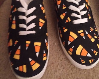May include: A pair of black canvas shoes with a white lace-up closure. The shoes are decorated with a pattern of orange and yellow candy corn.