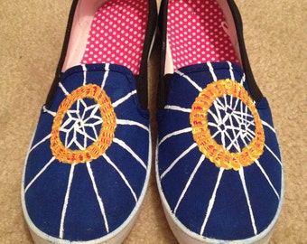 Könnte beinhalten: Ein Paar blaue Canvas-Slipper mit weißen Streifen und einem gelben und orangefarbenen Kreisdesign. Die Schuhe haben ein rosafarbenes Punktefutter.