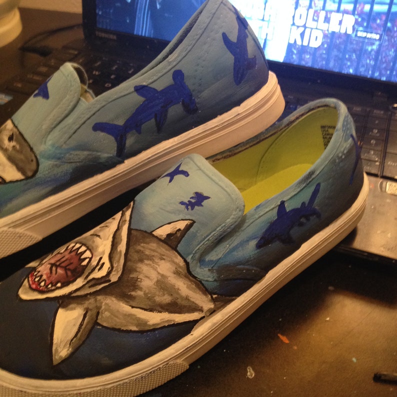 May include: A pair of blue canvas slip-on shoes hand-painted with a shark and other sea creatures. The shoes have a white sole and a yellow interior.